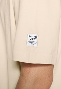 Manga de algodón beige con un parche blanco de Reebok que dice "Tinte Natural" cosido cerca del dobladillo en un antebrazo con pecas.