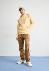 Beige hooded sweatshirt with front pocket, brown patterned joggers featuring white stripes, and green-accented white sneakers.