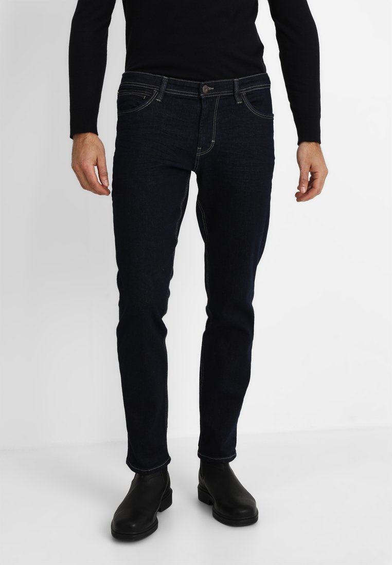 Dark blue denim jeans with a slim fit, featuring five pockets, subtle stitching details, and a classic button closure. Worn with black ankle boots.