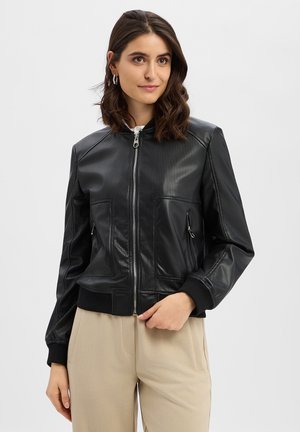 Woman with shoulder-length dark hair wearing a black perforated leather jacket and beige pants, standing against a plain white background.