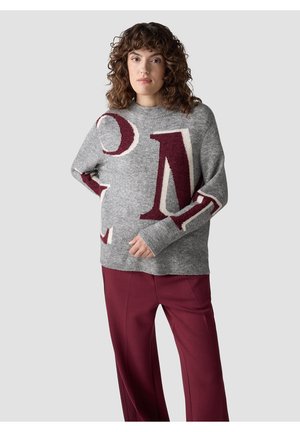 Woman with curly hair wearing a gray sweater with large maroon and white letters and maroon pants, standing against a plain background.