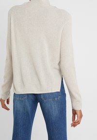 Persoon in een beige geribbelde trui met zijsplitten en een blauwe spijkerbroek, gezien vanaf de achterkant tegen een effen achtergrond.