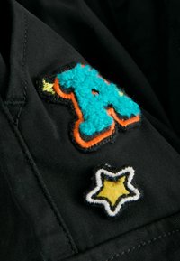 Tissu noir avec deux patchs : une lettre "A" turquoise duveteuse bordée d'orange et une étoile jaune avec un contour noir et blanc.