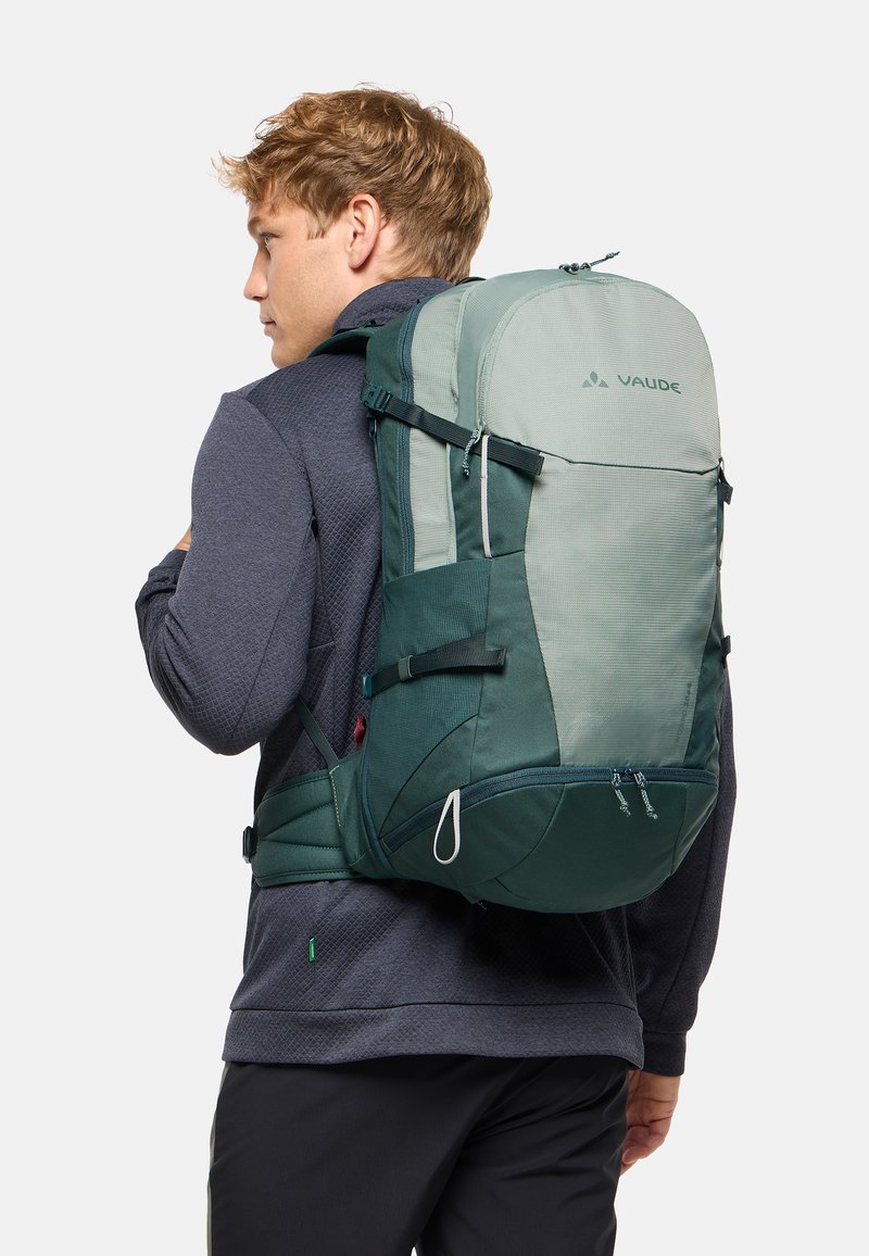 Joven con ropa oscura que lleva una gran mochila Vaude verde y gris, visto desde atrás y ligeramente de lado.