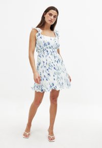 Robe à motifs floraux blancs, ornée d'accents bleus et verts, avec des bretelles volantes et une taille cintrée, se terminant par un ourlet évasé à bords en scallop.