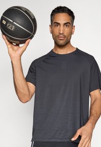 Μαύρο αθλητικό μπλουζάκι με υφασμένο μοτίβο διαβάθμισης, κοντά μανίκια και σχεδίαση ράγκλαν. Ένα μαύρο μπάσκετ κρατιέται σε ένα χέρι.