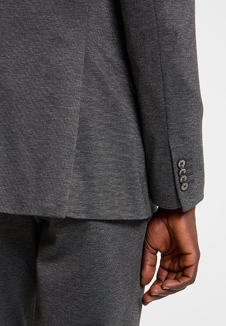 Traje gris con una tela texturizada. Primer plano de la manga que muestra cuatro acentos de botones y un ajuste hecho a medida. Construcción suave y elegante con un diseño moderno.