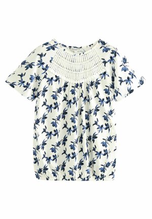 Blouse blanche à manches courtes avec motif floral bleu et large panneau en dentelle blanche au crochet sur le haut de la poitrine.