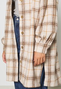 Persona che indossa un cappotto buttoned a quadri beige sopra una maglietta grigia e jeans blu, con una mano appoggiata di fianco e le maniche del cappotto raccolte ai polsi.