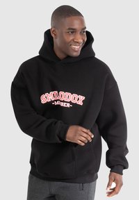 Černá mikina s kapucí, která má vpředu červenou a bílou grafiku, žebrované manžety a uvolněný střih. Vyrobena z měkké a pohodlné tkaniny.