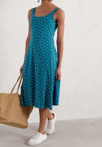 Teal klänning med vita prickar, rundad halsringning, figurnära liv, utsvängd kjol, i kombination med vita sneakers och en vävd väska.