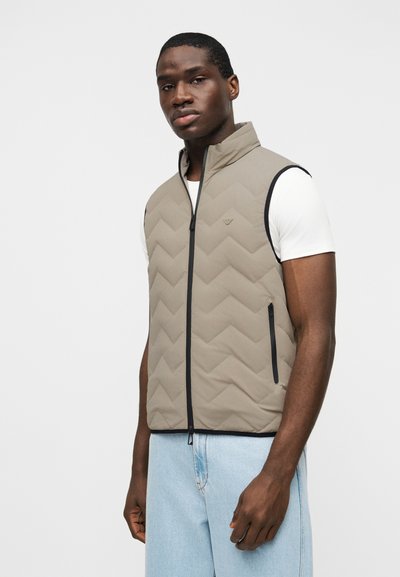 Άνδρας που φοράει ένα ανοιχτό καφέ παλτό χωρίς μανίκια με ύφασμα καπιτονέ πάνω από λευκό t-shirt και ανοιχτό μπλε τζιν, στέκεται μπροστά σε λευκό φόντο.