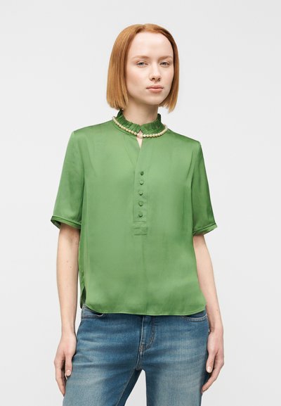 Πράσινη μεταξωτή μπλούζα με γιακά με βολάν, κοντά μανίκια και πατιλέτα με έξι κουμπιά. Συνδυάζεται με μπλε τζιν παντελόνι.