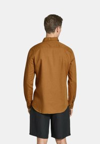 Camicia a maniche lunghe con colletto in lino marrone caldo, con bottoni sui polsini, orlo curvo e finitura testurizzata, indossata con pantaloni neri.