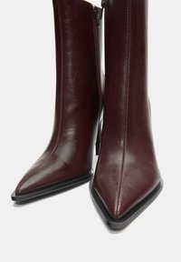 Burgunderrøde læderankelstøvler med spids tå, lynlås i siden og en slank sort hæl. Glat tekstur med synlige syningsdetaljer.