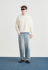 Νέος άνδρας που φορά γυαλιά ηλίου, μπεζ φούτερ Puma, ξεβάμμένα μπλε τζιν με μοτίβο λογότυπου Puma και μαύρα παπούτσια, στέκεται σε μπλε χαλί.