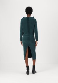 Gebreid jurk in diepgroen met een gestructureerde afwerking. Heeft een split aan de achterkant, een geribde zoom en lange mouwen. Gepaarde met zwarte enkellaarsjes.