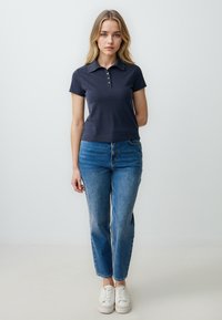 Polo shirt blu navy con colletto e tre bottoni, abbinato a jeans a gamba dritta di un leggero blu chiaro, leggermente sbiaditi e sneakers bianche.