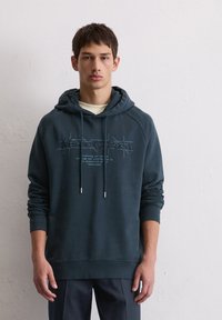 Haut à capuche bleu marine en mélange de coton, doté d'une capuche avec cordon de serrage, d'une broderie sur le devant et de poignets côtelés. Associé à un pantalon sombre.