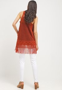 Rostfarbener, gestrickter Tanktop mit offenem Muster und Fransen am Saum. Kombiniert mit weißen, eng anliegenden Hosen und beigefarbenen Sandalen mit Absatz.