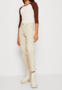 Krémové džíny rovného střihu v kombinaci s béžovým topem s dlouhými rukávy, který má hnědé raglánové rukávy, a robustními béžovými botami.