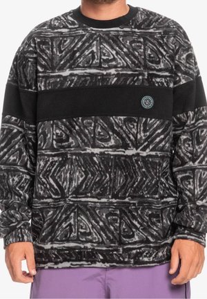 Mies, jolla on musta- ja harmaakuvioinen fleeceneule, jossa on leveä musta vaakaraita ja pieni pyöreä logo rinnassa, yhdistettynä violetteihin housuihin.