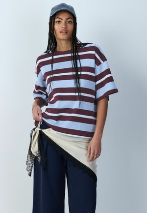 Kobieta z długimi warkoczami, nosząca niebieską czapkę, luźną koszulkę w niebiesko-bordowe paski, granatowe spodnie oraz kremowy szal z czarną koronką związany w talii.