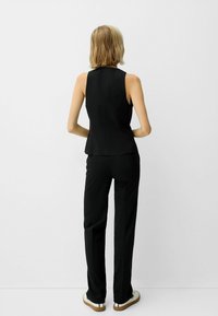 En blond kvinna står vänd mot en vit vägg i en ärmlös svart topp, svarta byxor och vita sneakers med gummisålar.