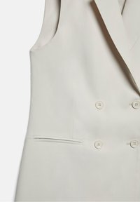 Beżowy blazer bez rękawów z podwójnymi guzikami i poziomą kieszenią po lewej stronie.