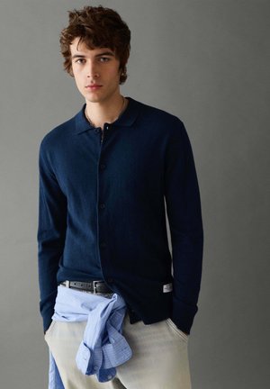 Junger Mann trägt einen dunkelblauen, geknöpften Cardigan und hellbeige Hosen mit einem hellblauen Hemd, das um die Taille gebunden ist, vor einem grauen Hintergrund.