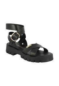 Sandal de piel negra con correa para el tobillo ajustable, tiras cruzadas y suela de goma gruesa y texturizada. Presenta un detalle de logotipo en tono dorado.
