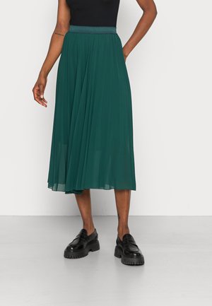 Faltenrock in Maxilänge in Dunkeltürkis, fließender Stoff, elastischer Taillenbund, kombiniert mit schwarzen Plateauschuhen. Glatte Textur und knielanges Design.