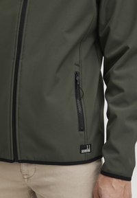 Olive green softshell jacket with a black zipper, side pocket, and logo patch. The fabric has a smooth texture with subtle patterns.