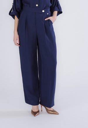 Femme portant un pantalon large bleu marine et un haut assorti avec des boutons dorés et des chaussures à talons hauts métalliques bronze, debout devant un fond uni.