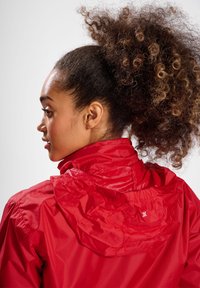 Junge Frau mit lockigem Haar, die eine rote Kapuzenjacke trägt, seitlich und von hinten vor einem schlichten Hintergrund gezeigt.