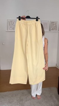 Lichtgele pantalons met wijde pijpen en een elastische tailleband, gemaakt van een soepele stof, weergegeven aan een hanger tegen een neutrale achtergrond.