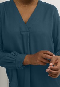 Blouse teal avec un col en V, manches longues se terminant par des poignets froncés et une texture douce. Deux poches avant sont visibles.