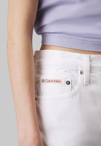 Jeans de mezclilla blanca con una etiqueta roja de Calvin Klein visible en el bolsillo trasero. El brazo está en un top recortado de color púrpura claro, mostrando piel.