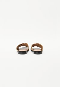 Sandales marron avec deux brides croisées, semelle noire plate, texture lisse et design minimaliste. Formes arrondies sur le dessus.
