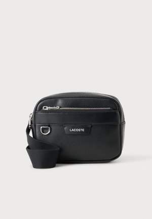 Sac bandoulière Lacoste en cuir noir texturé avec fermeture éclair argentée, poche avant, sangle réglable et logo sur le panneau avant sur fond blanc.