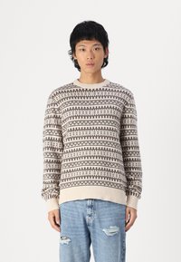 Suéter beige con estampado que presenta diseños geométricos en marrón, con puños y dobladillo acanalados, combinado con unos jeans desgastados de color azul claro.