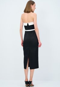 Abito nero di raso senza spalline con una fascia bianca sul busto, di lunghezza media con uno spacco sul retro e chiusura con cerniera. Indossato con scarpe nere con tacco alto.