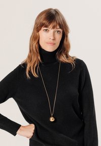 Schwarzer Rollkragensweater mit Rippbündchen, dazu eine lange goldene Anhängerkette mit einem runden Charm, vor einem neutralen Hintergrund präsentiert.