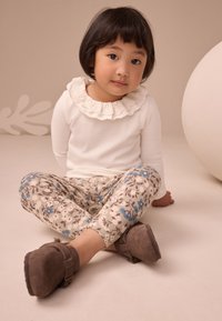 Maglia bianca a maniche lunghe con collo arricciato, pantaloni crema e marroni a fantasia floreale, e scarpe in camoscio marrone con dettaglio fibbia.