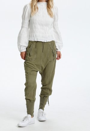 Vrouw draagt witte blouse met lange mouwen en geplooid, olijfgroene drop-crotch broek met grote voorzakken en witte sneakers, staand op witte achtergrond.