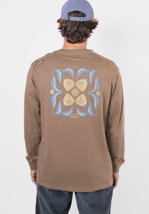 Personne portant une chemise marron à manches longues avec un motif floral abstrait bleu et beige et le texte "OXBOW" dans le dos, ainsi qu'une casquette bleu clair.