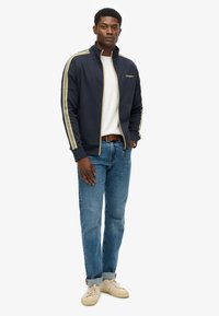 Marineblauw jack met rits en gouden streepaccenten, wit T-shirt, blauwe spijkerbroek en beige sneakers. Het jack heeft een hoge kraag en een merkdetail.