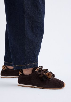 Persona con vaqueros azul oscuro y zapatillas de ante marrón oscuro con cordones con pompones y detalles con estampado de leopardo en los cuellos del talón.