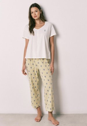 Kobieta stoi boso, ubrana w biały krótki top i żółte spodnie z małym niebieskim kwiatowym wzorem.