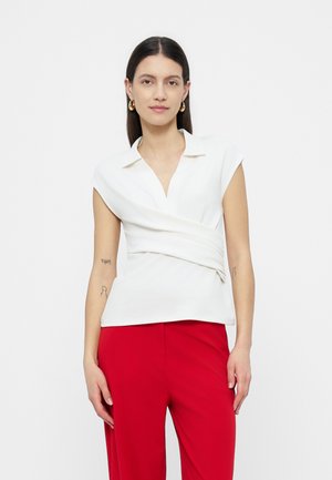 Γυναίκα με λευκό κοντομάνικο τοπ τύπου wrap με γιακά και κόκκινο παντελόνι, στέκεται με τα χέρια χαλαρά σε απλότερο φόντο.